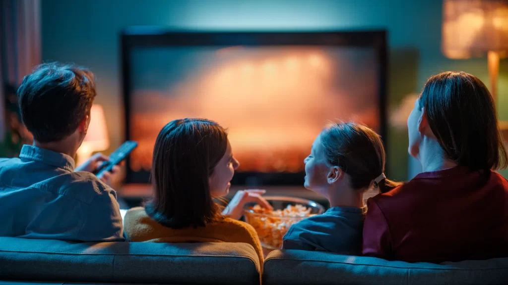 Family watching digitized home videos on TV together – Your Photo Assistant Family of four watching home videos together on a large screen television in a dark cozy living room