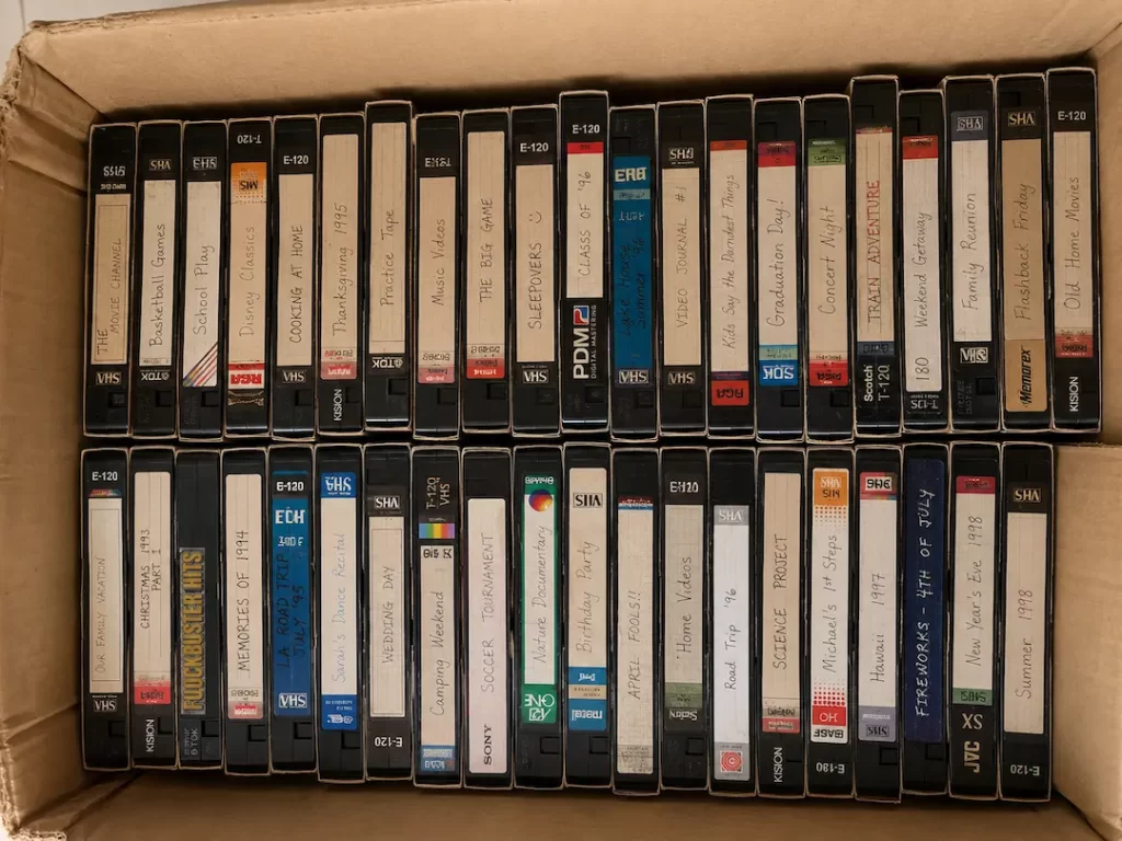 Box of family VHS home video tapes with handwritten labels – Your Photo Assistant Cardboard box packed with labeled VHS home video tapes from the 1990s including Christmas, wedding day, and vacation recordings