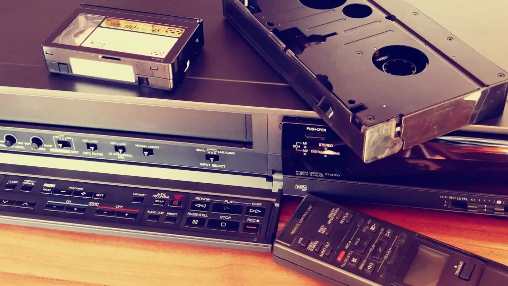 Vintage VCR player with VHS tapes — home video playback equipment – Your Photo Assistant Vintage VCR player with VHS cassette tapes stacked on top and a remote control on a wooden surface